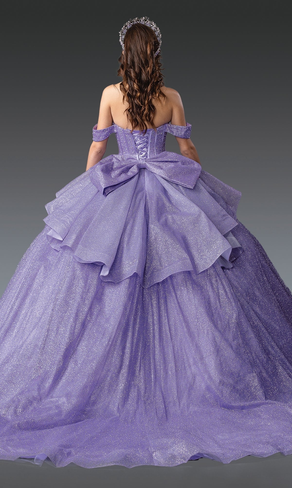A woman with long brown hair models the Quinceanera Dress 1937 By Dancing Queen, a sparkling lavender ball gown with layered tulle, off-the-shoulder straps, and a large back bow, set against a gray background.